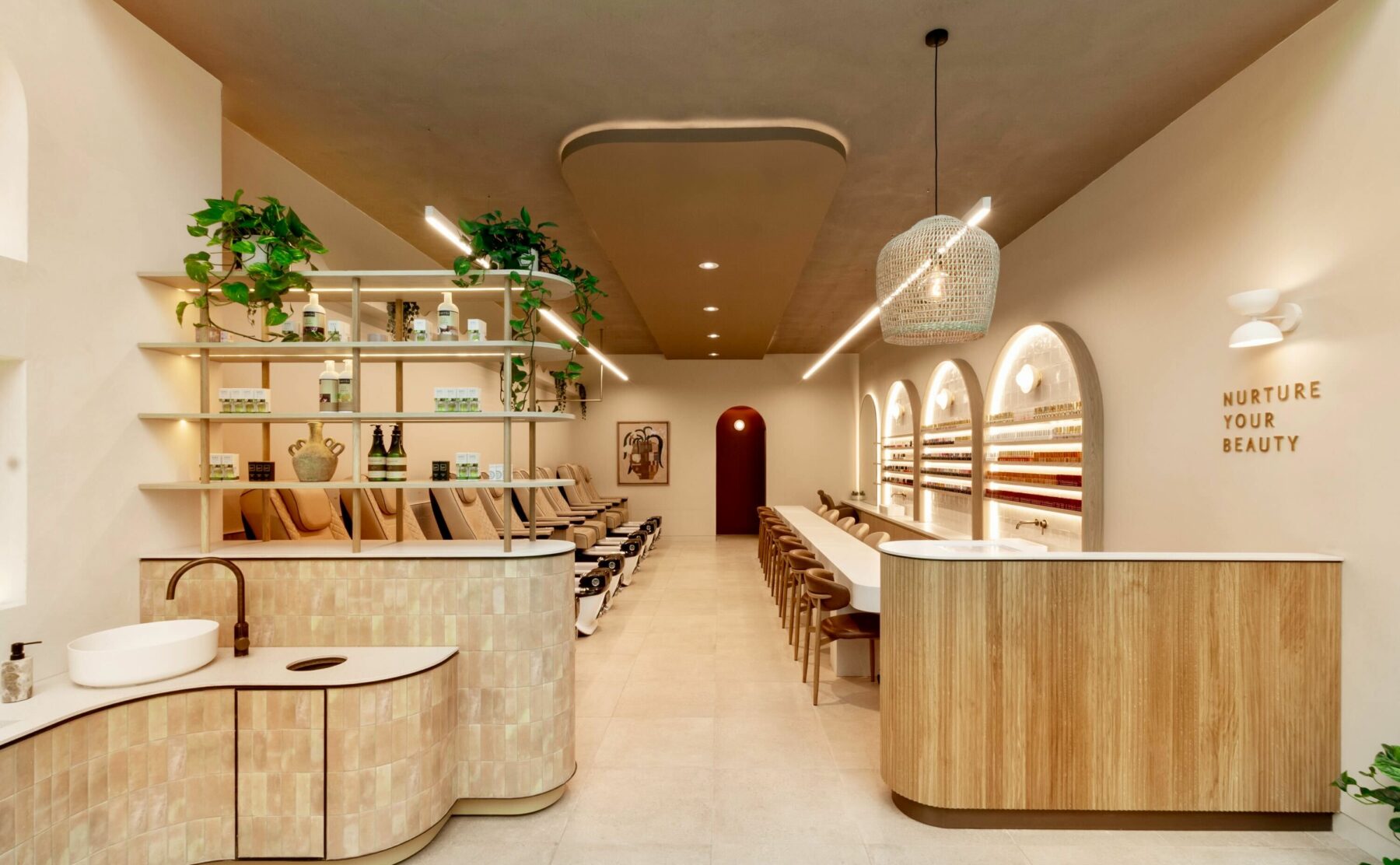 The spa’s inviting front view highlights a soothing neutral palette with fluted timber, textured tiles, and layered shelving. Pedicure stations line the left, manicure stations the right, while soft lighting and greenery create a calming, wellness‑driven ambience.