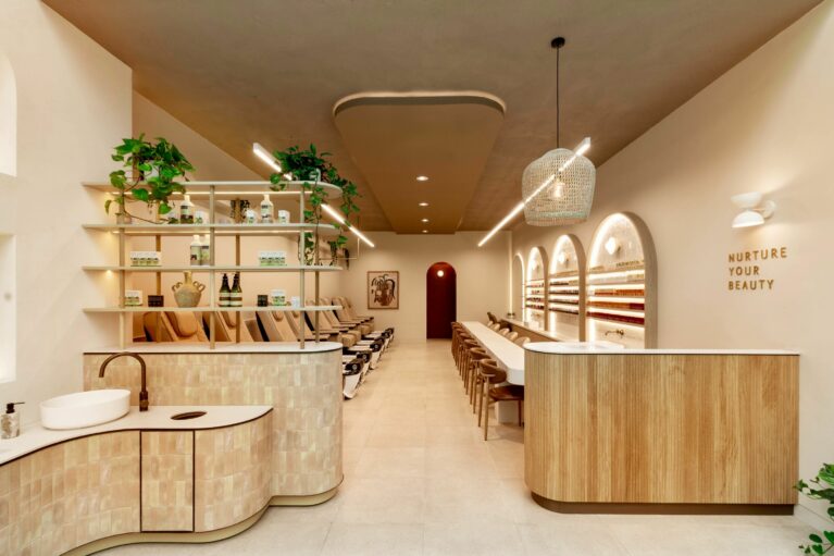 The spa’s inviting front view highlights a soothing neutral palette with fluted timber, textured tiles, and layered shelving. Pedicure stations line the left, manicure stations the right, while soft lighting and greenery create a calming, wellness‑driven ambience.