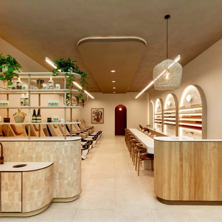 The spa’s inviting front view highlights a soothing neutral palette with fluted timber, textured tiles, and layered shelving. Pedicure stations line the left, manicure stations the right, while soft lighting and greenery create a calming, wellness‑driven ambience.
