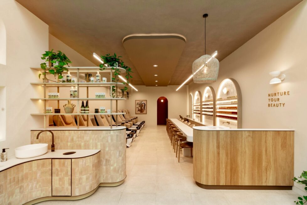 The spa’s inviting front view highlights a soothing neutral palette with fluted timber, textured tiles, and layered shelving. Pedicure stations line the left, manicure stations the right, while soft lighting and greenery create a calming, wellness‑driven ambience.