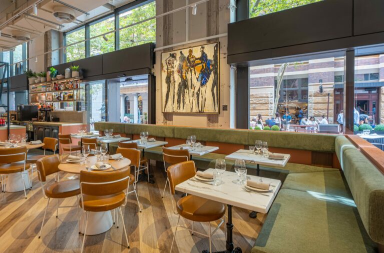 Margot Osteria's dining area features a soft palette, fresh finishes, and refined textures. Olive green banquette seating wraps the interior with leather-upholstered chairs and sleek stone tabletops, offering comfort and contrast against the building’s raw structural shell.
