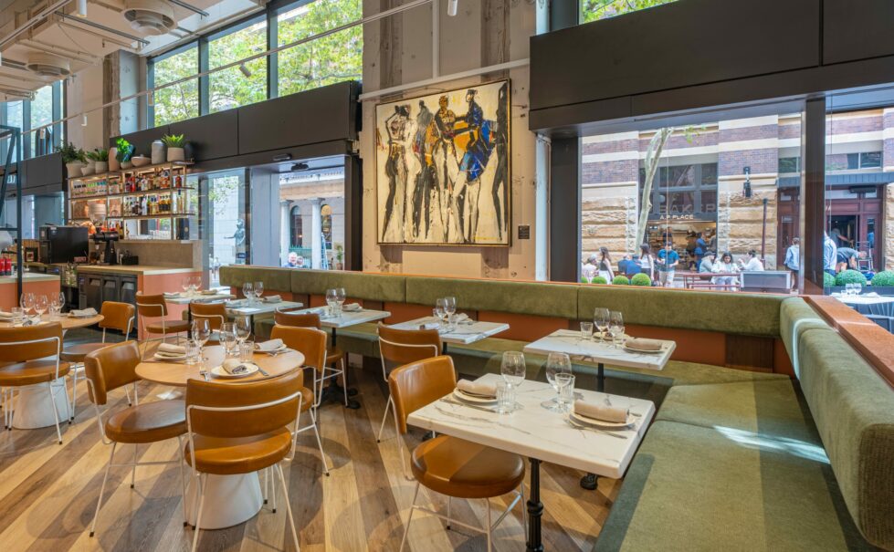 Margot Osteria's dining area features a soft palette, fresh finishes, and refined textures. Olive green banquette seating wraps the interior with leather-upholstered chairs and sleek stone tabletops, offering comfort and contrast against the building’s raw structural shell.