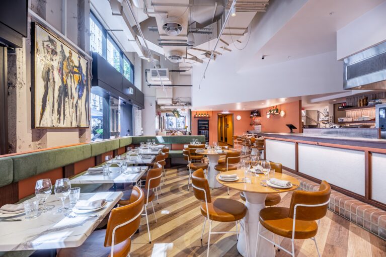 Margot Osteria's dining area features a soft palette, fresh finishes, and refined textures. Olive green banquette seating wraps the interior with leather-upholstered chairs and sleek stone tabletops, offering comfort and contrast against the building’s raw structural shell.