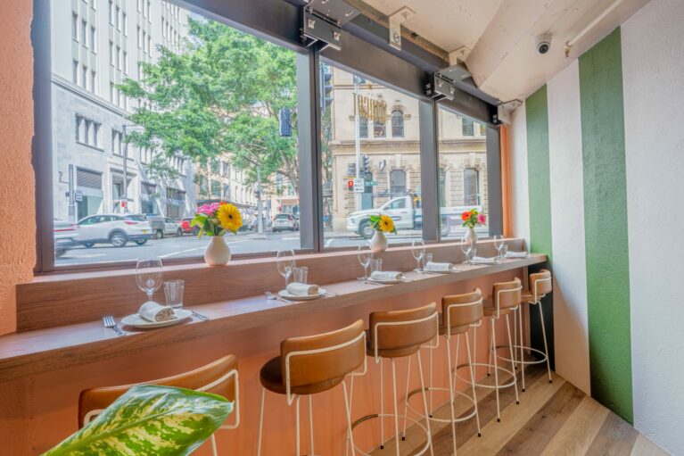 A vibrant mix of urban charm and Italian ease, this counter-height seating along Margot Osteria’s large street-facing windows invites guests to dine in rhythm with the city. Tanned leather bar stools line a timber counter, softened by playful florals and crisp settings that balance casual sophistication with visual warmth.