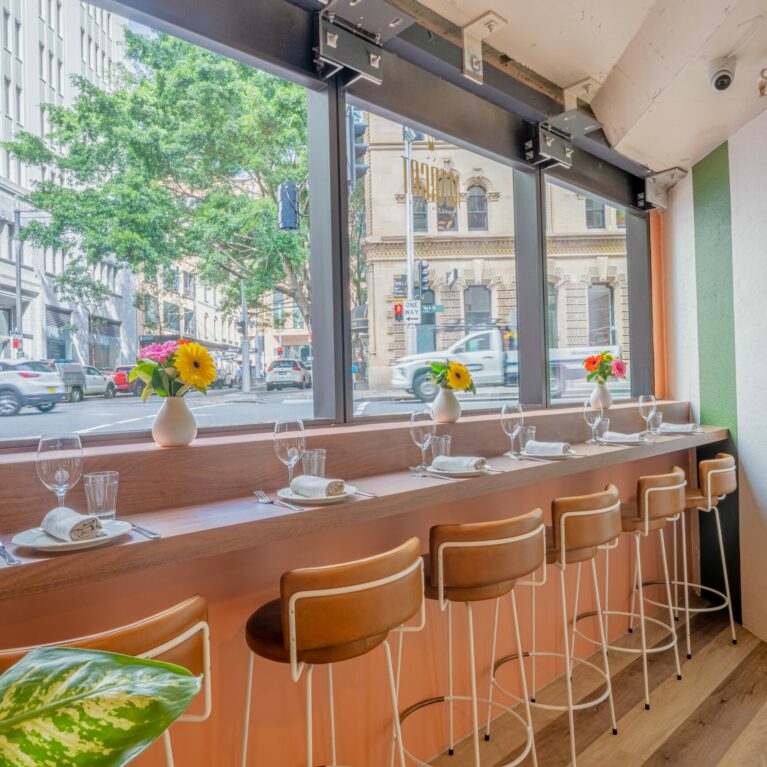 A vibrant mix of urban charm and Italian ease, this counter-height seating along Margot Osteria’s large street-facing windows invites guests to dine in rhythm with the city. Tanned leather bar stools line a timber counter, softened by playful florals and crisp settings that balance casual sophistication with visual warmth.