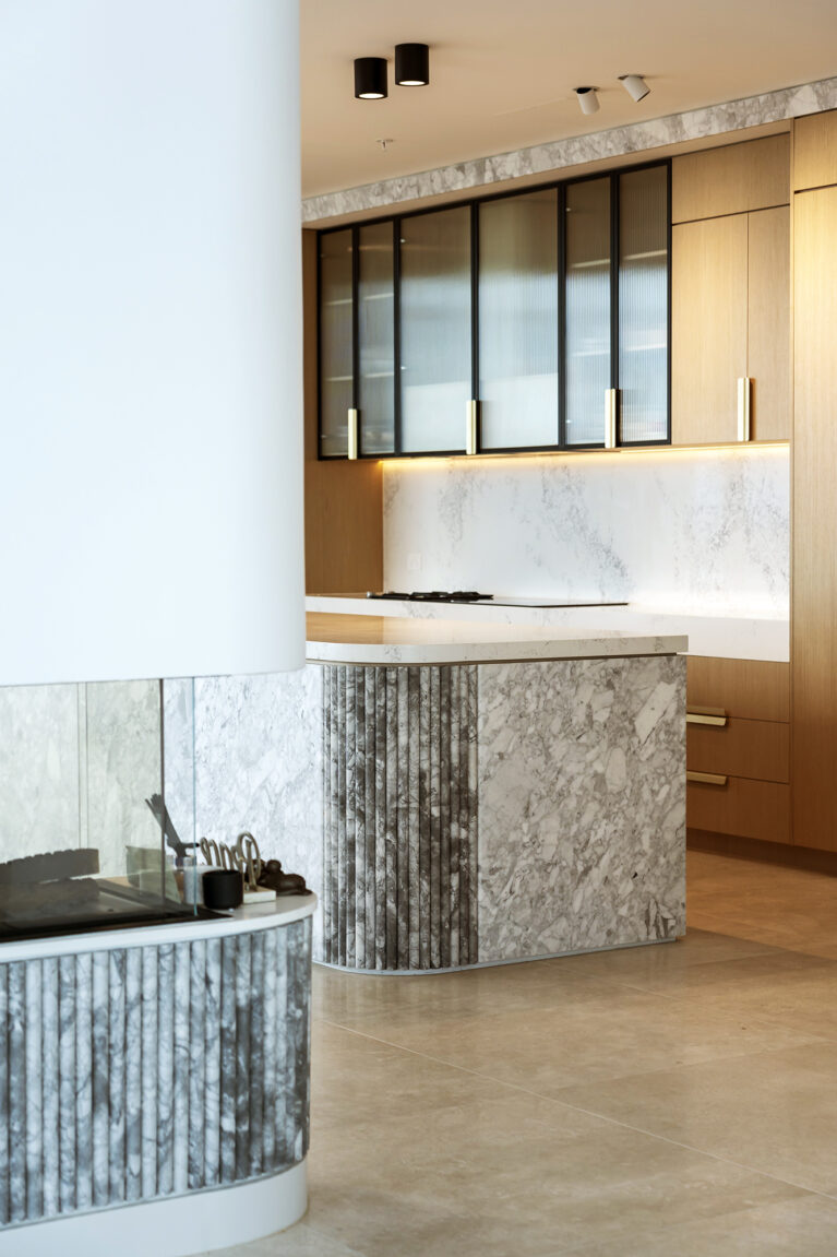 A view of the kitchen and adjoining fireplace area highlights the harmonious blend of Calacatta marble, light oak cabinetry, and fluted detailing, creating a seamless flow between functional and relaxation spaces.