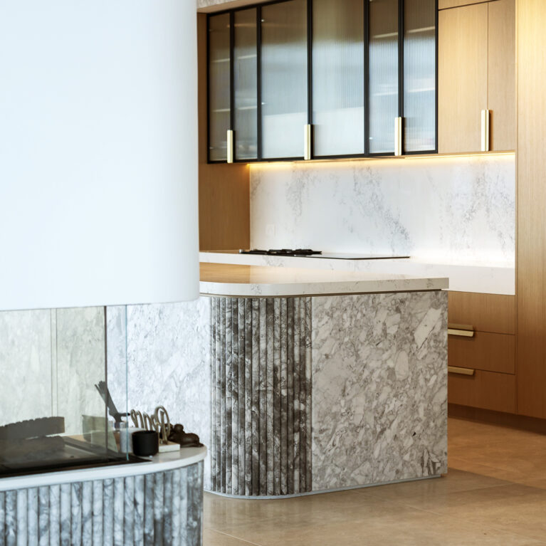 A view of the kitchen and adjoining fireplace area highlights the harmonious blend of Calacatta marble, light oak cabinetry, and fluted detailing, creating a seamless flow between functional and relaxation spaces.