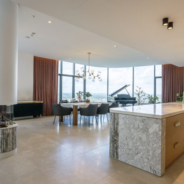 The open-plan kitchen and dining area, framed by floor-to-ceiling windows, showcases Calacatta marble and oak finishes, enhancing the natural light and expansive views. The design invites seamless transitions between cooking, dining, and entertaining.