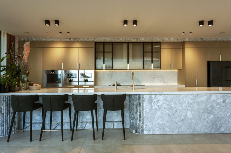 The kitchen exudes luxury and functionality, featuring a Calacatta marble island and light oak cabinetry. Black accents and integrated appliances enhance the modern aesthetic, while soft lighting highlights the refined textures of the materials.