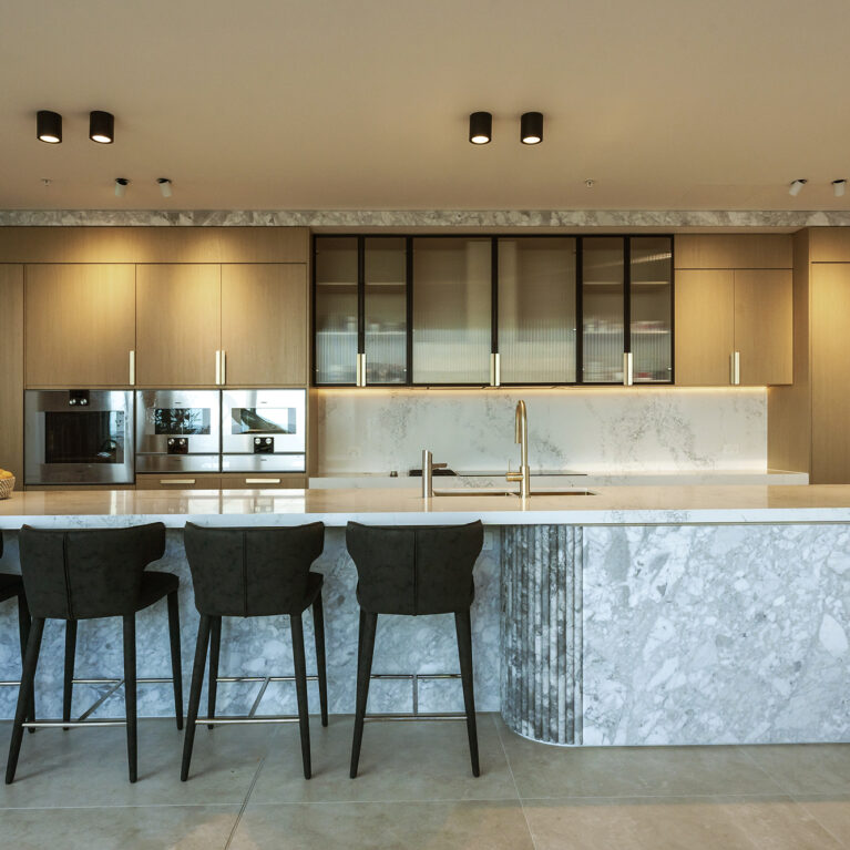 The kitchen exudes luxury and functionality, featuring a Calacatta marble island and light oak cabinetry. Black accents and integrated appliances enhance the modern aesthetic, while soft lighting highlights the refined textures of the materials.