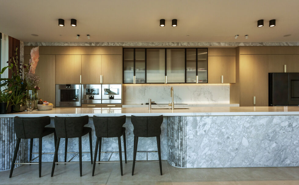 The kitchen exudes luxury and functionality, featuring a Calacatta marble island and light oak cabinetry. Black accents and integrated appliances enhance the modern aesthetic, while soft lighting highlights the refined textures of the materials.