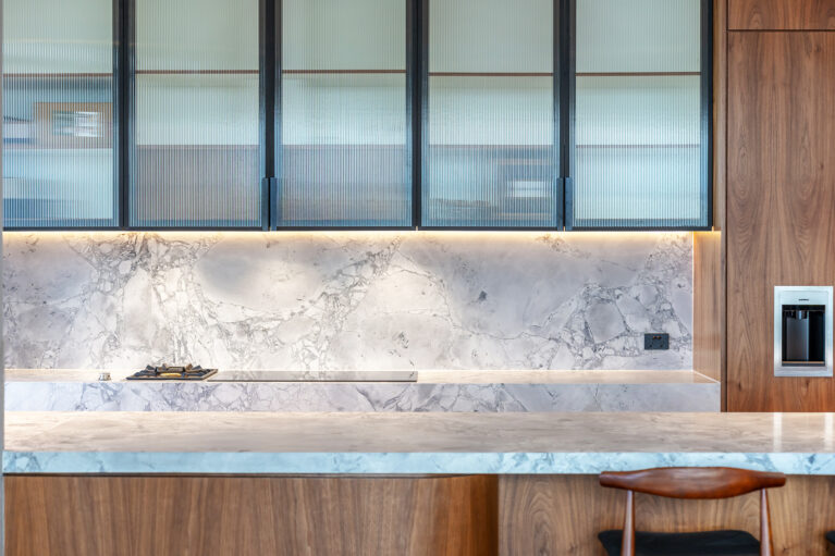 A refined interplay of textures and lighting brings depth to the kitchen, showcasing honed marble, fluted glass cabinetry, and warm wood.