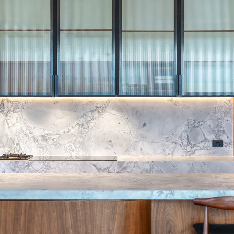 A refined interplay of textures and lighting brings depth to the kitchen, showcasing honed marble, fluted glass cabinetry, and warm wood.