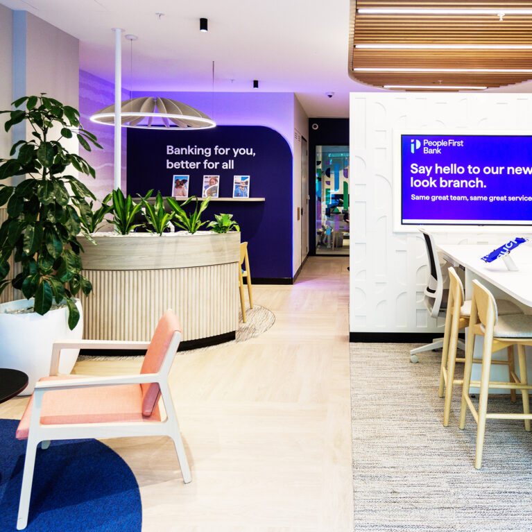 A bright and inviting lounge greets visitors, showcasing People First Bank's modern, community-focused ethos. This space features soft pastel tones, greenery, natural wood textures, and a mission statement wall that emphasizes the bank’s customer-centric philosophy.