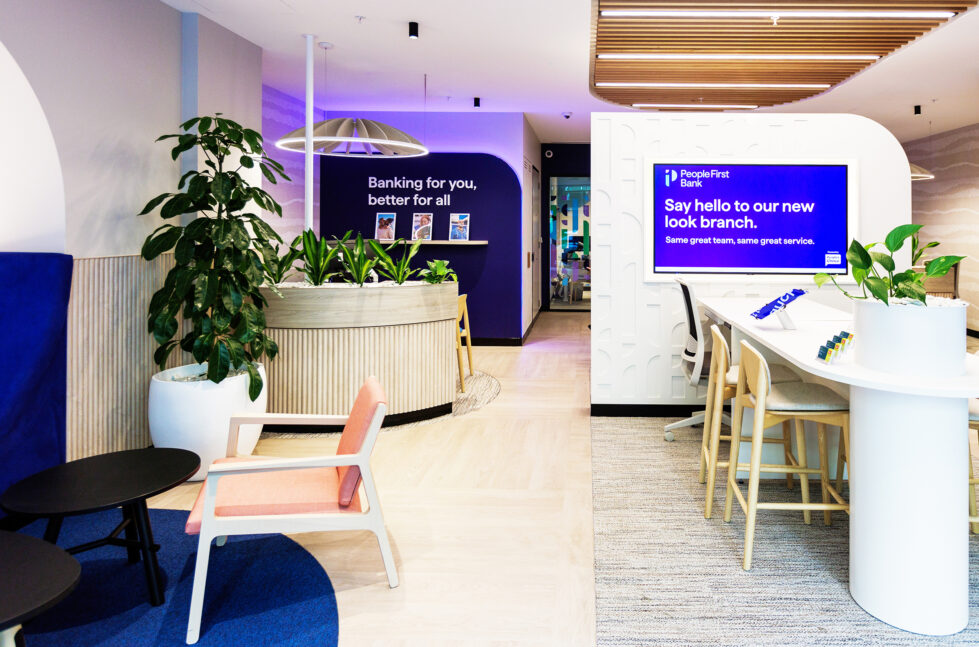 A bright and inviting lounge greets visitors, showcasing People First Bank's modern, community-focused ethos. This space features soft pastel tones, greenery, natural wood textures, and a mission statement wall that emphasizes the bank’s customer-centric philosophy.