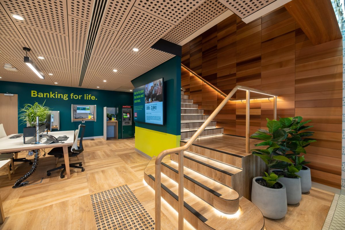 The connecting staircase at People’s Choice flagship branch, featuring warm wood tones, accent lighting, and lush greenery, creates an inviting transition between floors.