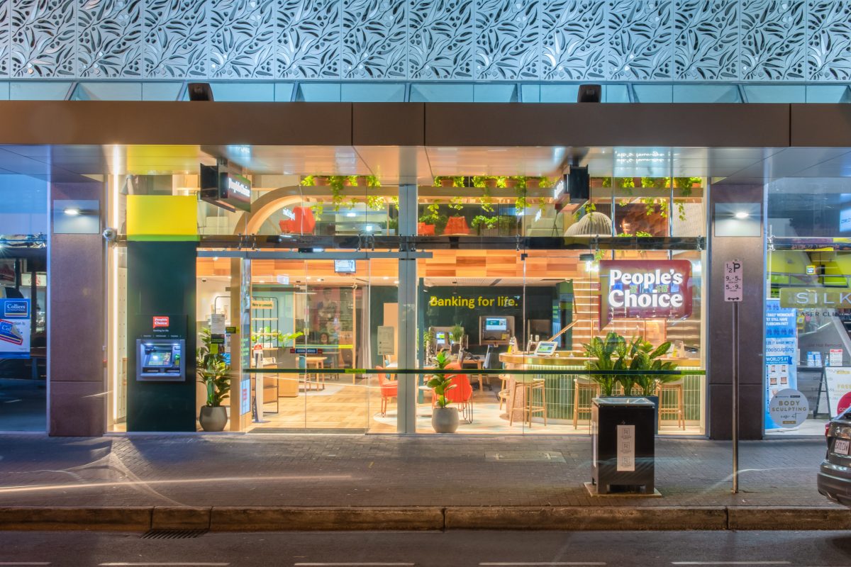 The People's Choice flagship branch storefront, warmly illuminated at night, showcases a modern, inviting interior with open seating, greenery, and a casual, customer-friendly design.