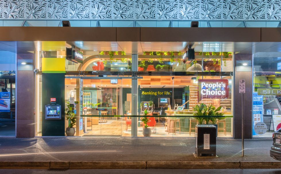 The People's Choice flagship branch storefront, warmly illuminated at night, showcases a modern, inviting interior with open seating, greenery, and a casual, customer-friendly design.