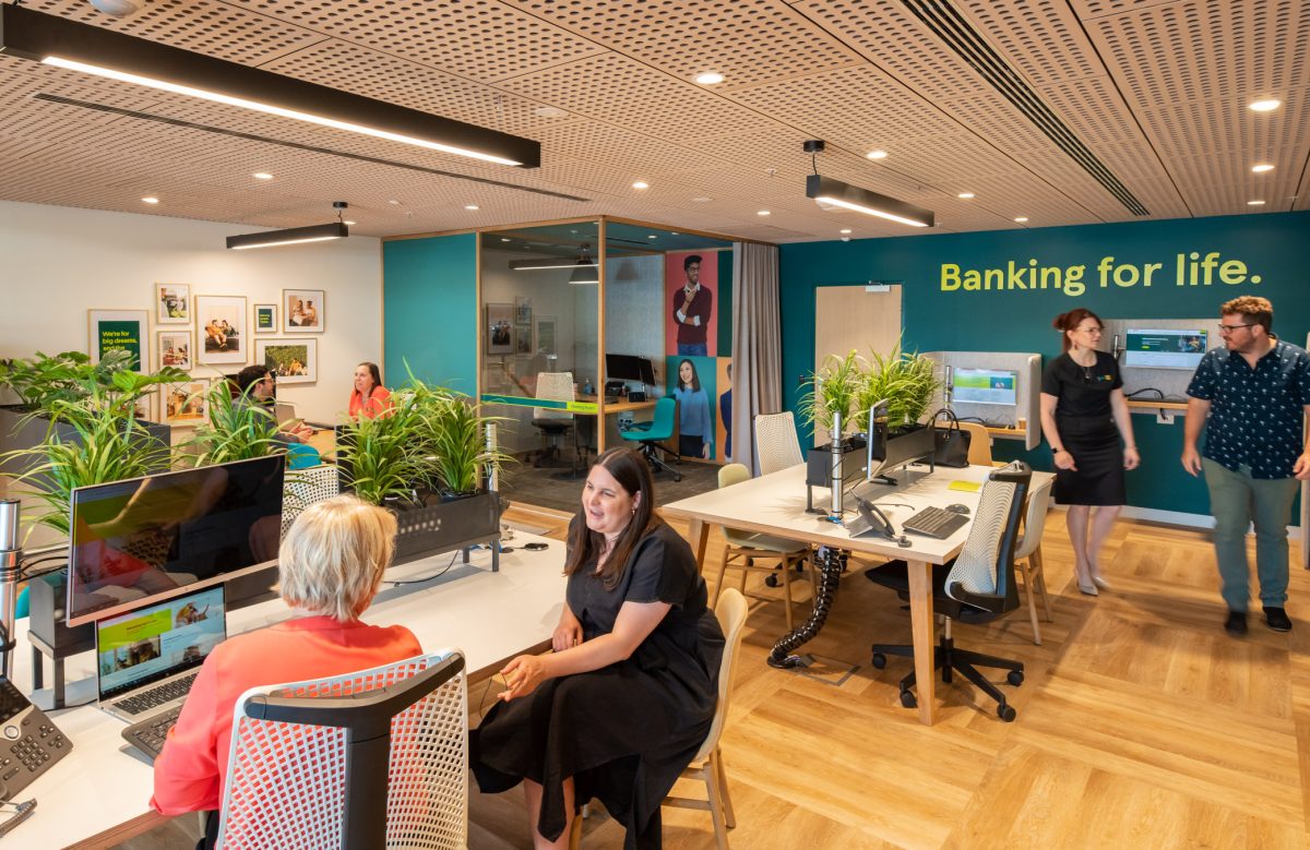 An open, collaborative workspace on the lower level of People’s Choice bank, featuring warm wood floors, vibrant teal accents, and abundant greenery.