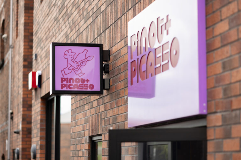 A bold purple sign pops against the rustic brick façade, creating a modern contrast. The illuminated hanging sign and 3D lettering add depth and visibility, setting the tone for a stylish and creative space inside.