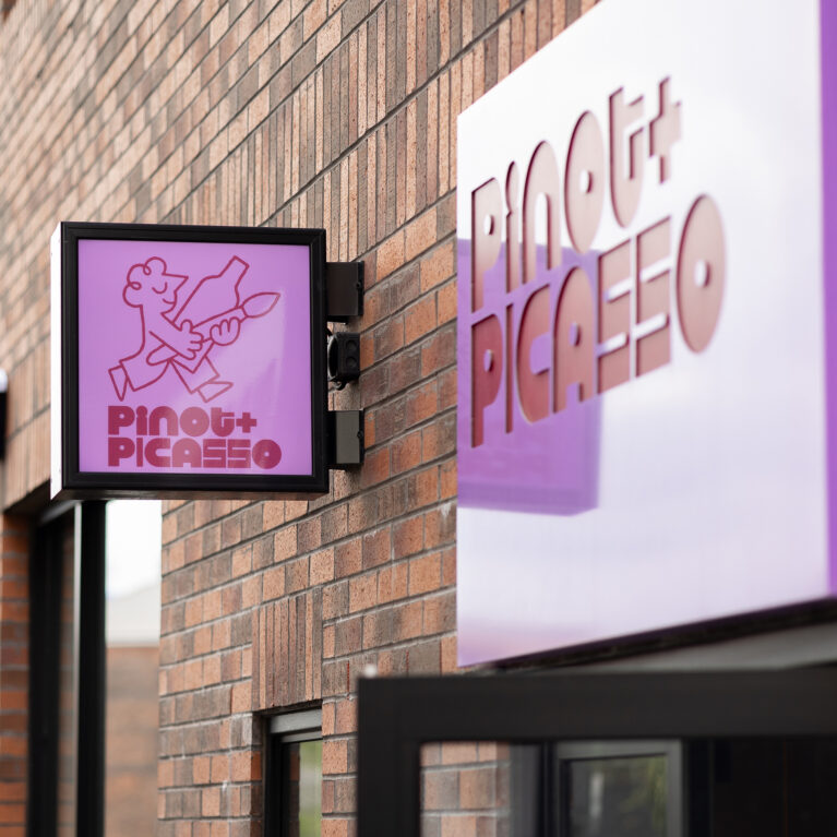 A bold purple sign pops against the rustic brick façade, creating a modern contrast. The illuminated hanging sign and 3D lettering add depth and visibility, setting the tone for a stylish and creative space inside.