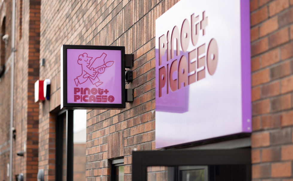 A bold purple sign pops against the rustic brick façade, creating a modern contrast. The illuminated hanging sign and 3D lettering add depth and visibility, setting the tone for a stylish and creative space inside.