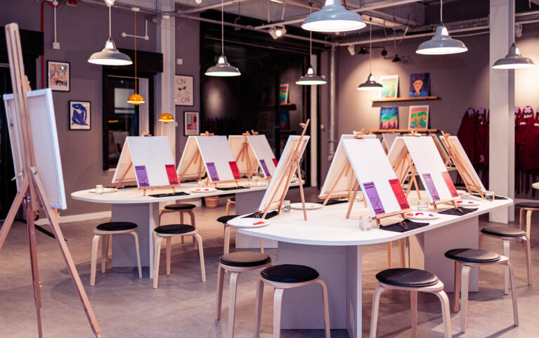 The open studio layout invites creativity, featuring curved tables with easels, simple stools, and industrial lighting. Neutral walls showcase vibrant artwork, while the polished floor and subtle color palette create a modern yet welcoming environment for fun group painting sessions.