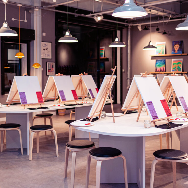 The open studio layout invites creativity, featuring curved tables with easels, simple stools, and industrial lighting. Neutral walls showcase vibrant artwork, while the polished floor and subtle color palette create a modern yet welcoming environment for fun group painting sessions.