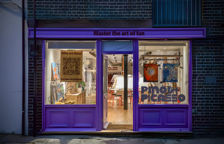 The inviting storefront glows with energy, drawing in guests with its bold purple accents and warm interior lighting. Playful typography and quirky signage set the stage for a creative space where art meets fun in a vibrant, social setting.