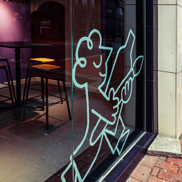 The storefront’s bold glass design features an oversized illustration that playfully hints at the creative experience inside. Reflected city architecture blends with the interior’s warm lighting, high stools, and deep-toned walls, creating a seamless connection between the studio and its urban surroundings.