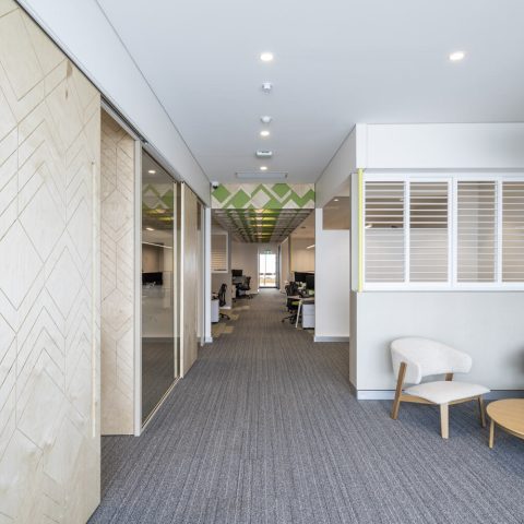 Corporate breakout furniture, white plantation shutters, acoustic felt lighting, sliding timber doors, glazed office partitions, commercial linear layout, geometric ceiling acoustic baffles, grey carpet tiles.