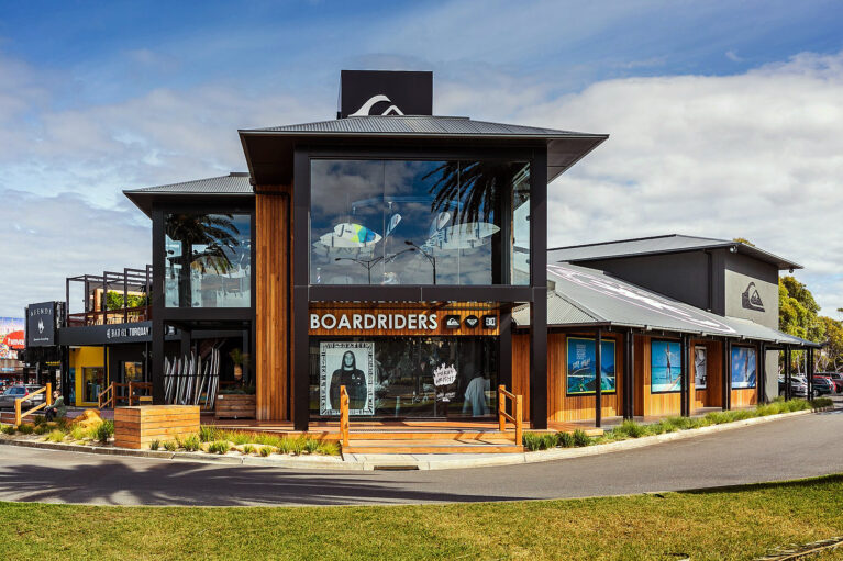 This vibrant exterior highlights the open, surf-inspired design. A glass façade frames suspended surfboards, while warm timber cladding complements the coastal surroundings. The inviting deck and detailed storefront underscore the space’s dual role as retail and community-focused.