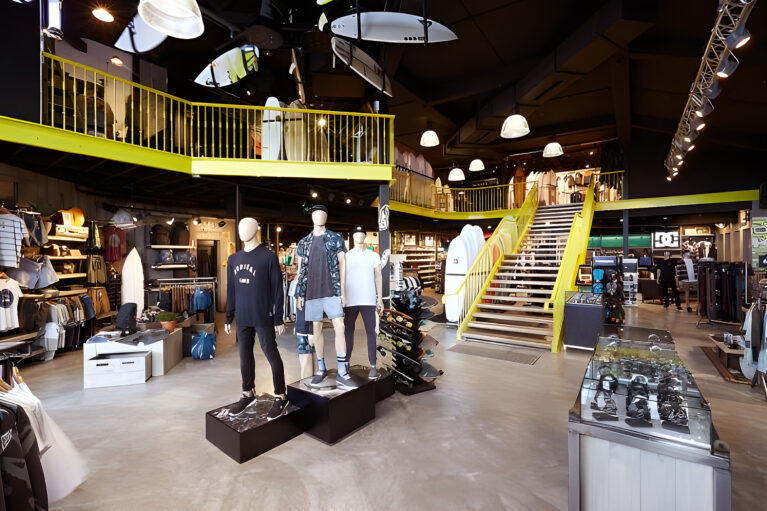 The vibrant interior features a multi-level layout with surfboards suspended overhead and mannequins showcasing apparel. Bold yellow accents on the staircase and balcony bring energy to the space, while curated product displays enhance the immersive shopping experience.