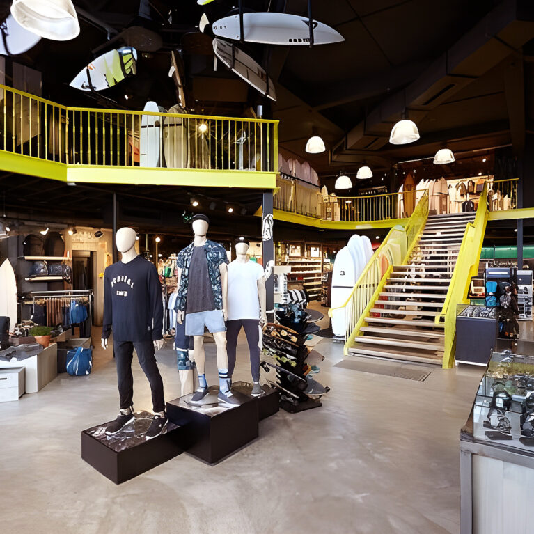 The vibrant interior features a multi-level layout with surfboards suspended overhead and mannequins showcasing apparel. Bold yellow accents on the staircase and balcony bring energy to the space, while curated product displays enhance the immersive shopping experience.