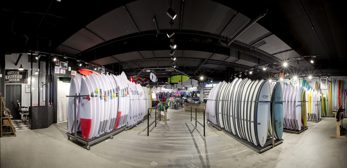 Open plan retail layout, industrial track lighting, custom surfboard racks, timber plank flooring, exposed matte black ceiling, commercial showroom design.