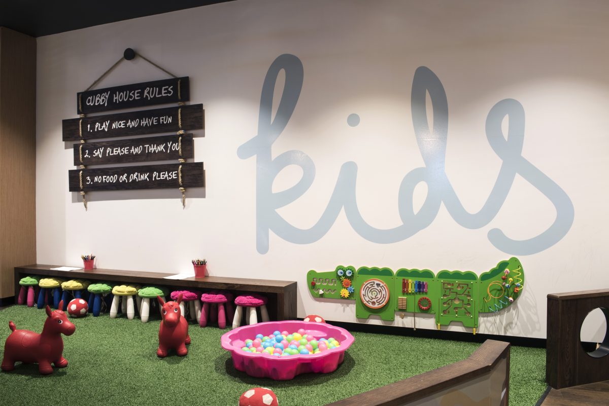 White retail wall graphics, artificial grass flooring, low timber activity benches, colorful kids plastic stools, crocodile wall panel toy, sensory ball pit pool, hanging timber signboard.