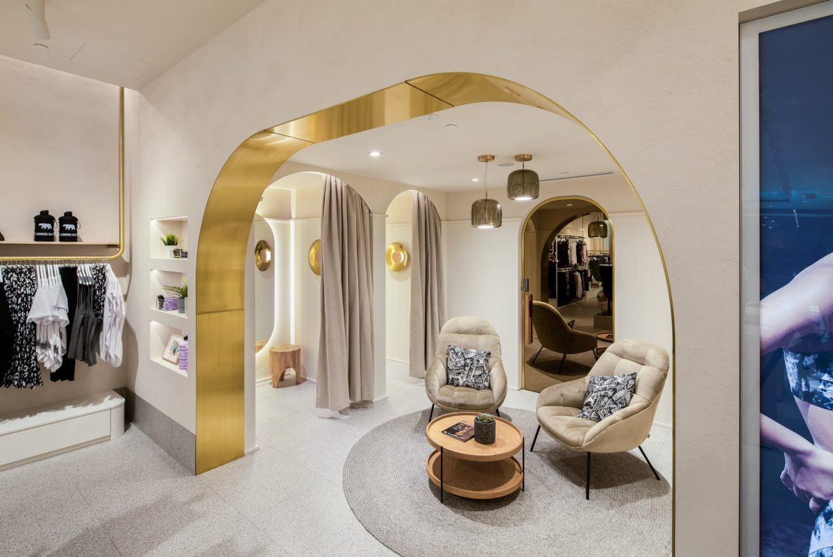 brushed brass portal arch, luxury fitting room suite, curved architectural threshold, terrazzo flooring, warm ambient cove lighting, coastal retail design, plush lounge seating, boutique retail fit-out