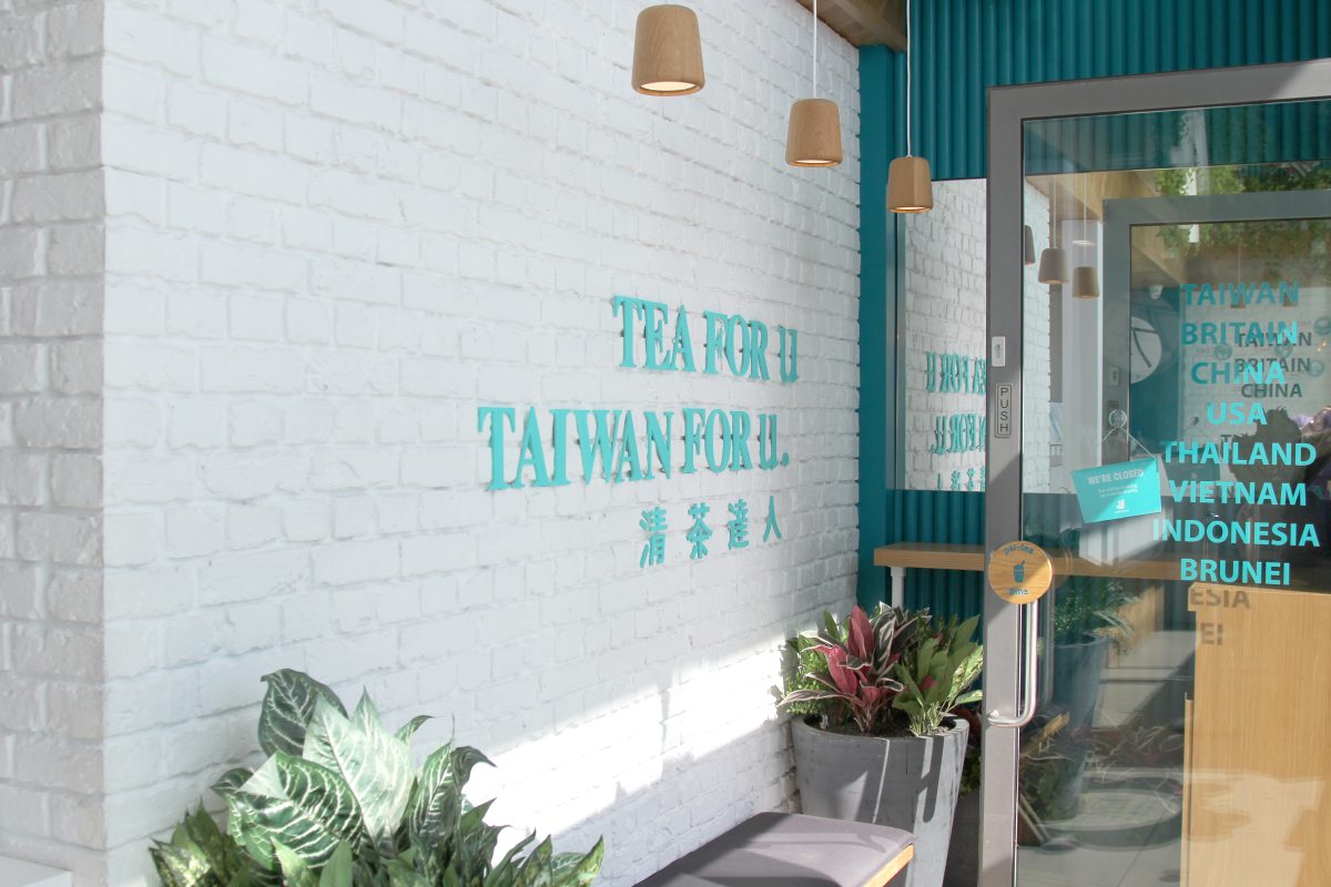 Painted white brick wall, teal corporate lettering, timber pendant lighting, gray fiberglass planter pots, commercial glass entryway, ribbed wall cladding.