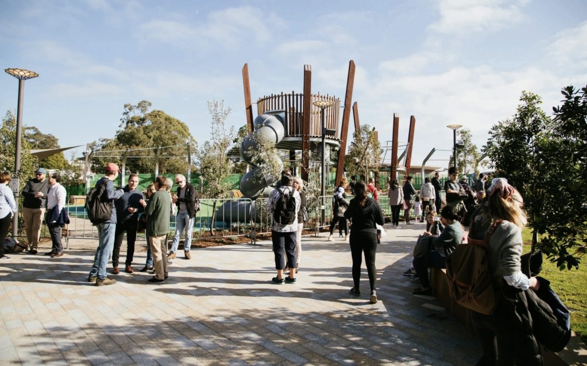A community playground with a modern, wooden structure, set within a sunny, tree-lined park.