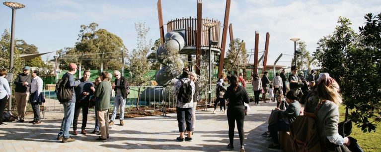 A community playground with a modern, wooden structure, set within a sunny, tree-lined park.