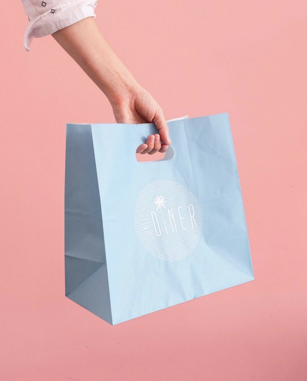 A stylish light-blue shopping bag with "DINER" branding and a palm tree graphic, held against a pastel pink background.