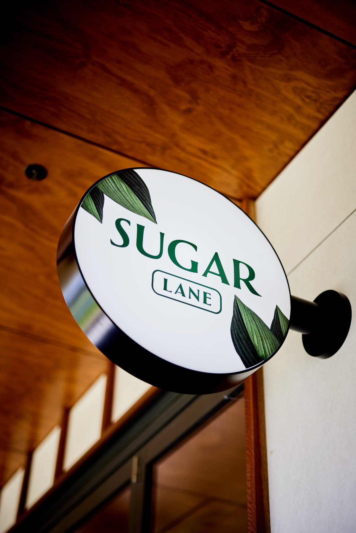 The Sugarlane blade sign, featuring a simple, elegant design with green foliage details on a white circular background.