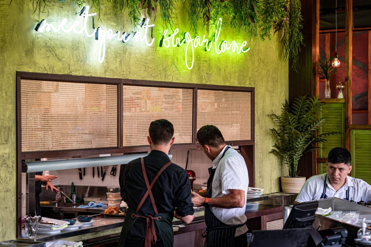 The back bar at Sugarlane, featuring a neon sign, tropical greenery, and warm wood accents.