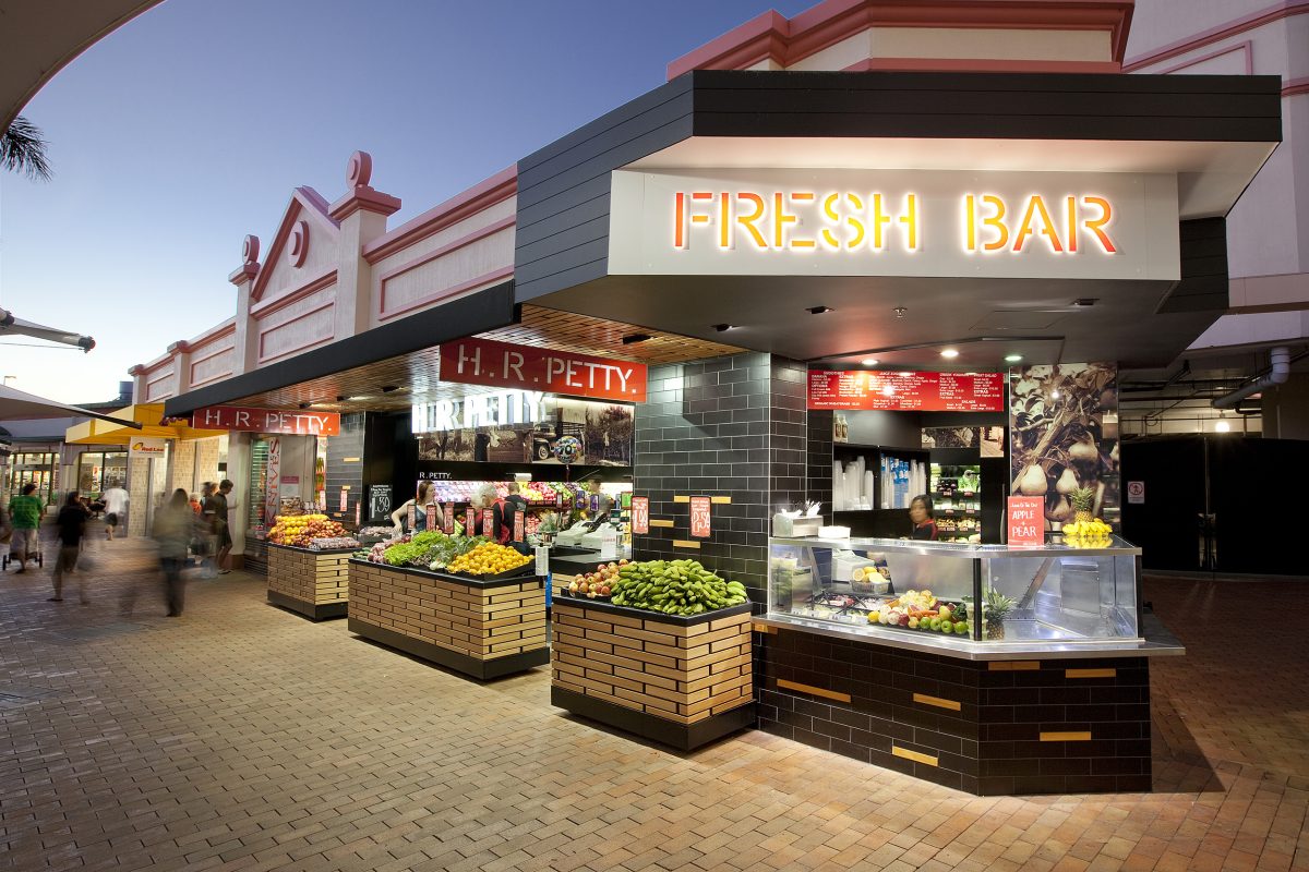 Shopping mall storefront, architectural corner canopy, neon style signage, refrigerated display case, outdoor market bins, brick tile pavement, open air retail concourse.