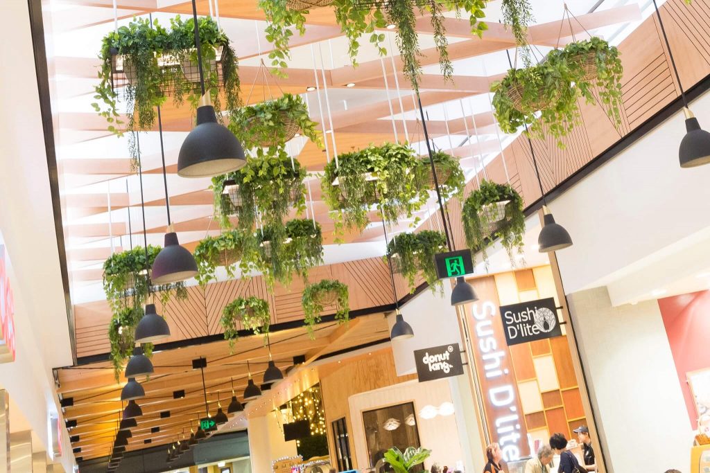 Biophilic mall design, suspended hanging planters, industrial dome pendants, atrium skylight, timber wall paneling, retail food precinct, vertical spatial layering.