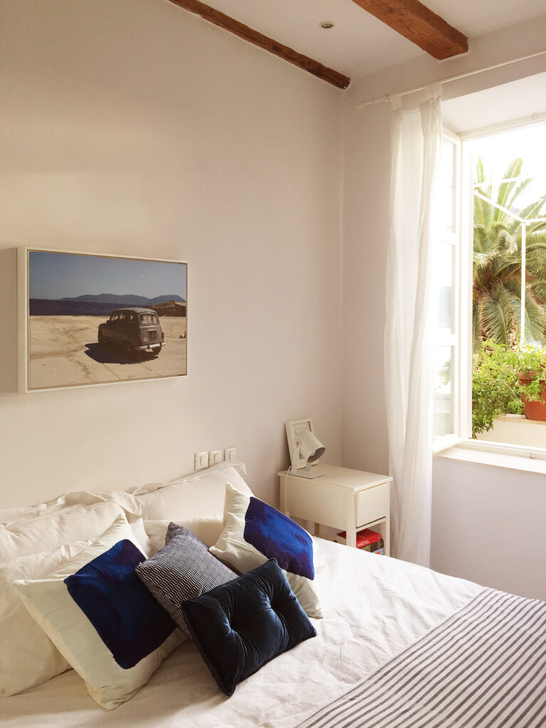 A serene bedroom with crisp white walls, wooden beams, and a bright open window framing lush greenery. Soft textures and artistic accents create an inviting retreat for relaxation.