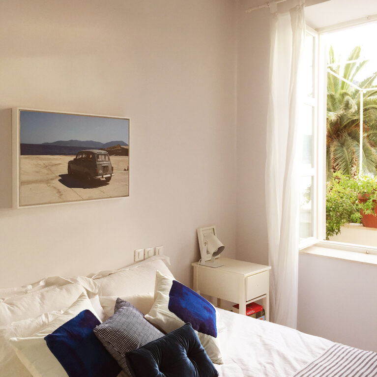 A serene bedroom with crisp white walls, wooden beams, and a bright open window framing lush greenery. Soft textures and artistic accents create an inviting retreat for relaxation.