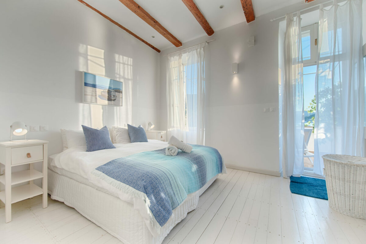 A serene bedroom with crisp white walls, wooden beams, and a bright open window framing lush greenery. Soft textures and artistic accents create an inviting retreat for relaxation.