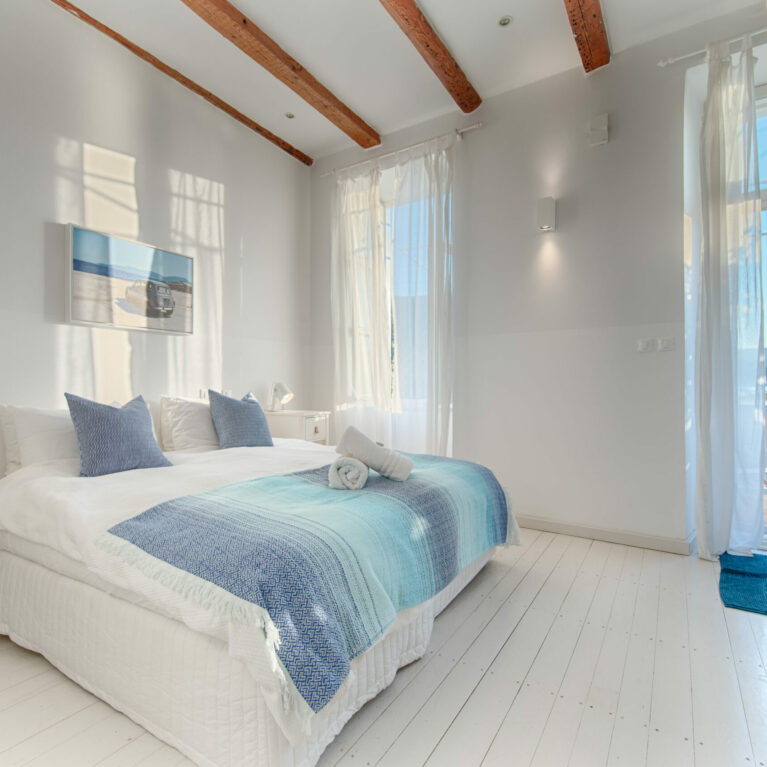 A serene bedroom with crisp white walls, wooden beams, and a bright open window framing lush greenery. Soft textures and artistic accents create an inviting retreat for relaxation.