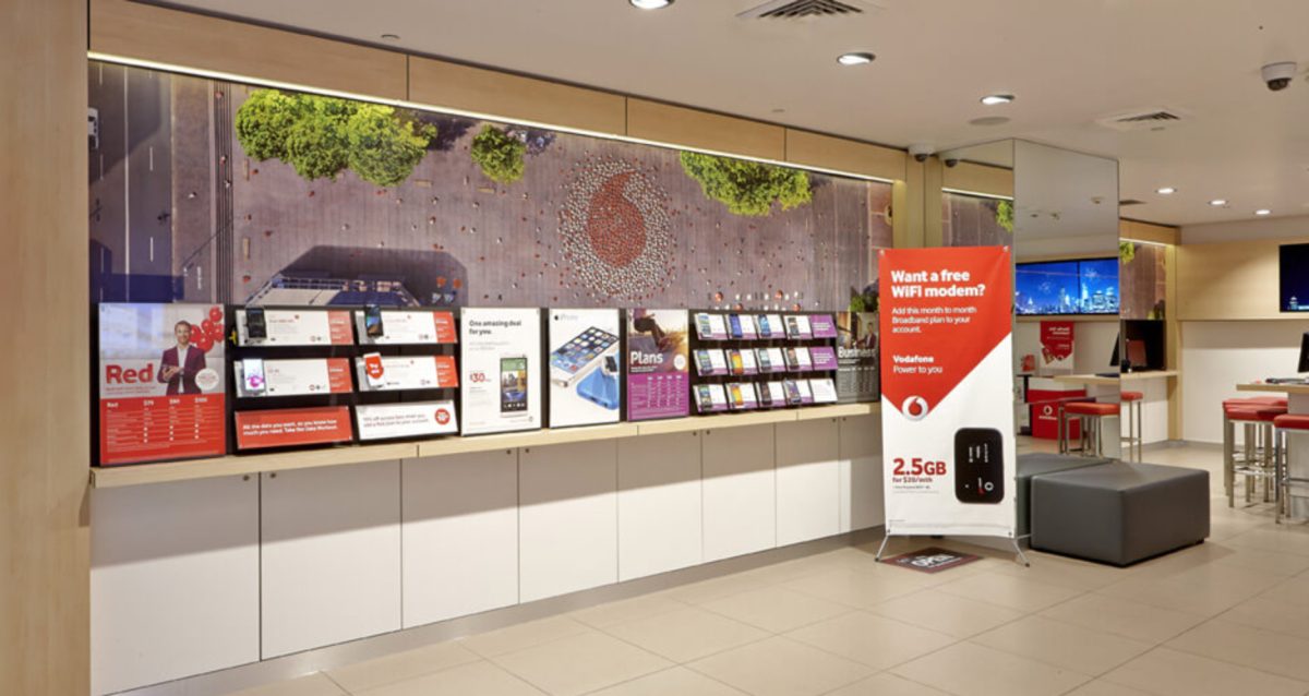 The product display wall in a Vodafone store, featuring organized plan information, smartphone displays, and a vibrant cityscape graphic.